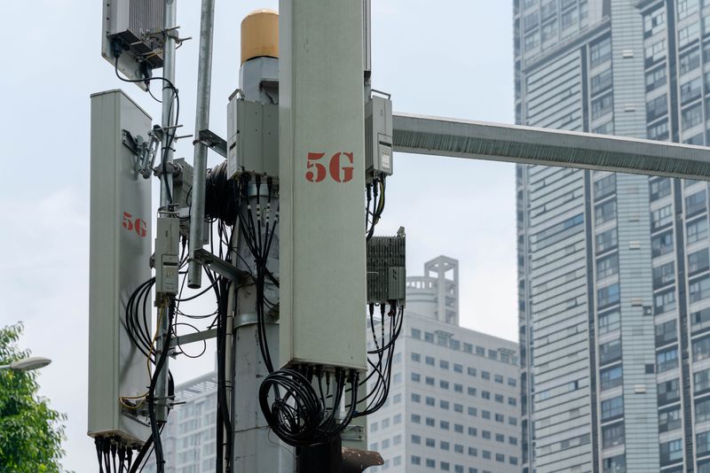 A close up photo of a 5G cellular network antenna with large urban building in the background. "5G" is visible on the side of the device, written in red. The antenna has wires visible and is attached to a central frame. 