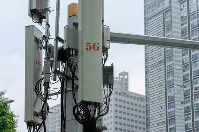 A close up photo of a 5G cellular network antenna with large urban building in the background. "5G" is visible on the side of the device, written in red. The antenna has wires visible and is attached to a central frame. 