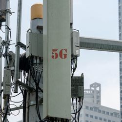 A close up photo of a 5G cellular network antenna with large urban building in the background. "5G" is visible on the side of the device, written in red. The antenna has wires visible and is attached to a central frame. 