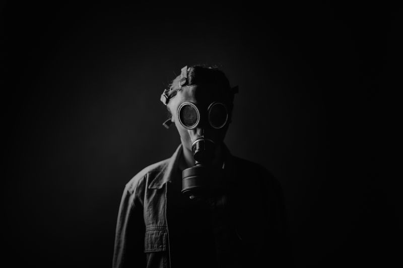 A black and white photo of someone wearing a gasmask an what looks like a denim coat. The person is standing against a black backdrop and only their right hand side has been lit, so they are partially obscured. 