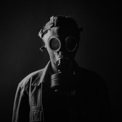 A black and white photo of someone wearing a gasmask an what looks like a denim coat. The person is standing against a black backdrop and only their right hand side has been lit, so they are partially obscured. 