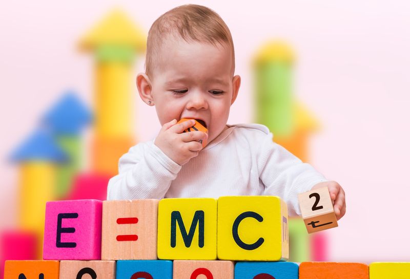 A photo of a baby plying with wooden bricks that spell out "E=MC2". The Child is placing the 2 brick on the end of the sequence and has another brick in its hand which it is about to chew on. Little genius. 
