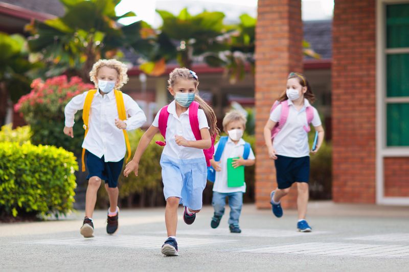 four young children running out of a school or kindergarten wearing face masks and differently coloured backpacks