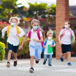 four young children running out of a school or kindergarten wearing face masks and differently coloured backpacks
