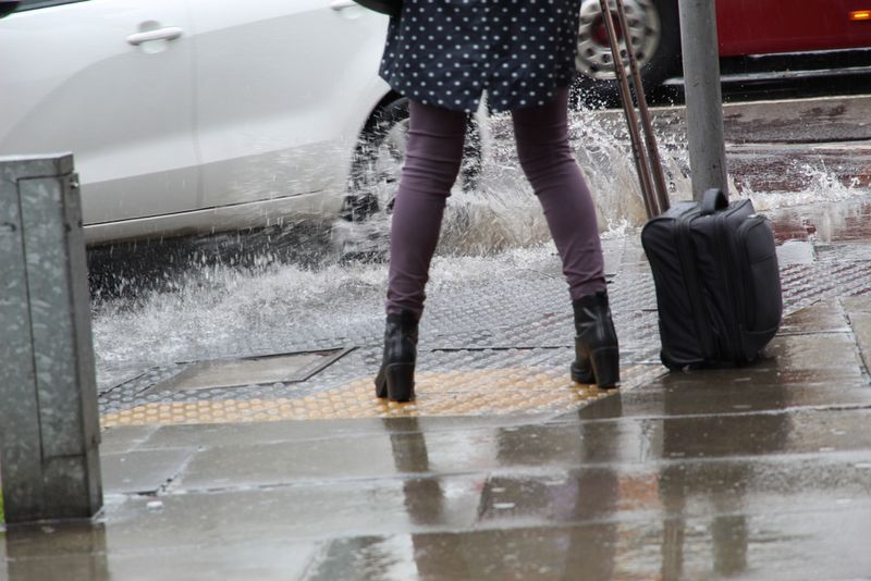 A photo showing the lower half of a woman wearing a white on black poke dot coat pulling a suitcase in the rain as she dodges splash from a passing car. She's is approaching the side of the pavement/sidewalk as the white car turns and sends water her way. 