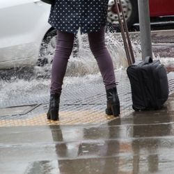 A photo showing the lower half of a woman wearing a white on black poke dot coat pulling a suitcase in the rain as she dodges splash from a passing car. She's is approaching the side of the pavement/sidewalk as the white car turns and sends water her way. 