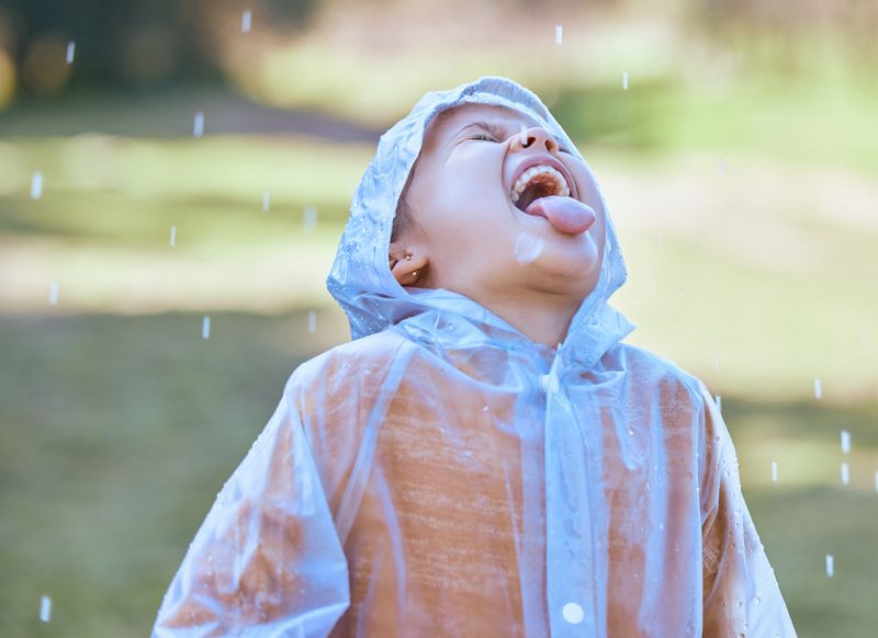 kid standing outside wearing a raincoat with mouth open and tongue out, trying to drink the rain