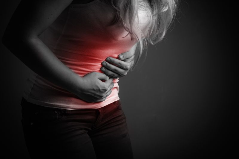 black and white photo of a woman with long hair holding her abdomen and bending forward as if in pain; there is red colouration over the abdomen to indicate pain; the woman is only visible between the shoulders and knees