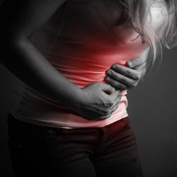 black and white photo of a woman with long hair holding her abdomen and bending forward as if in pain; there is red colouration over the abdomen to indicate pain; the woman is only visible between the shoulders and knees
