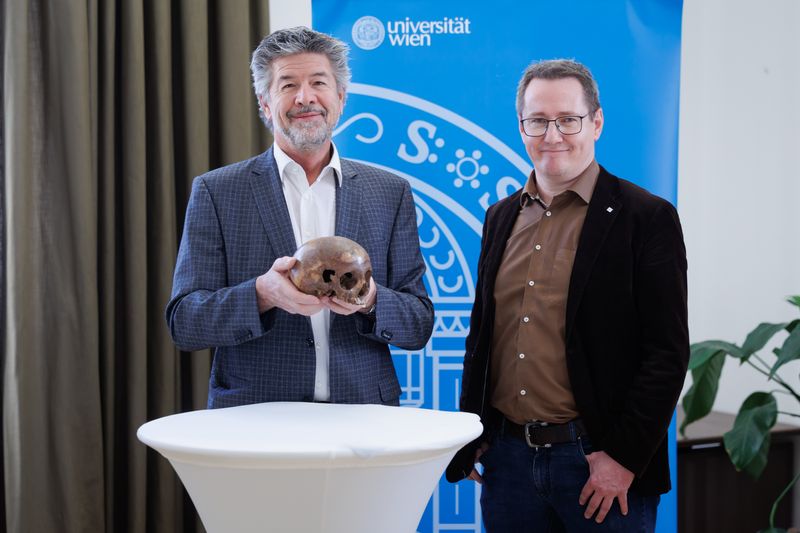 The photo shows the skull in the hands of Gerhard Weber who is standing next to Martin Steskal at a press conference. Weber is wearing a grew blazer and white shirt. He has a grey neat facial hear. Steskal is wearing a black blazer and a brown shirt. They are both standing in front of a small white stand.