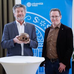 The photo shows the skull in the hands of Gerhard Weber who is standing next to Martin Steskal at a press conference. Weber is wearing a grew blazer and white shirt. He has a grey neat facial hear. Steskal is wearing a black blazer and a brown shirt. They are both standing in front of a small white stand.