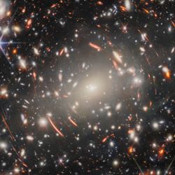A field of galaxies in space, dominated by an enormous, bright-white elliptical galaxy that is the core of a massive galaxy cluster. Many other elliptical galaxies can be seen around it. Also around it are short, curved, glowing red lines, which are images of distant background galaxies magnified and warped by gravitational lensing. A couple of foreground stars appear large and bright with long spikes around them.