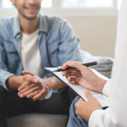 The photo shows a young man sat on a couch talking to a female doctor. The image is cropped so that the top of the man's head and his lower legs are out of frame. He is also blurry. In the right of the foreground, the arms and a folded leg of a female doctor can be seen holding a clipboard and a pen.