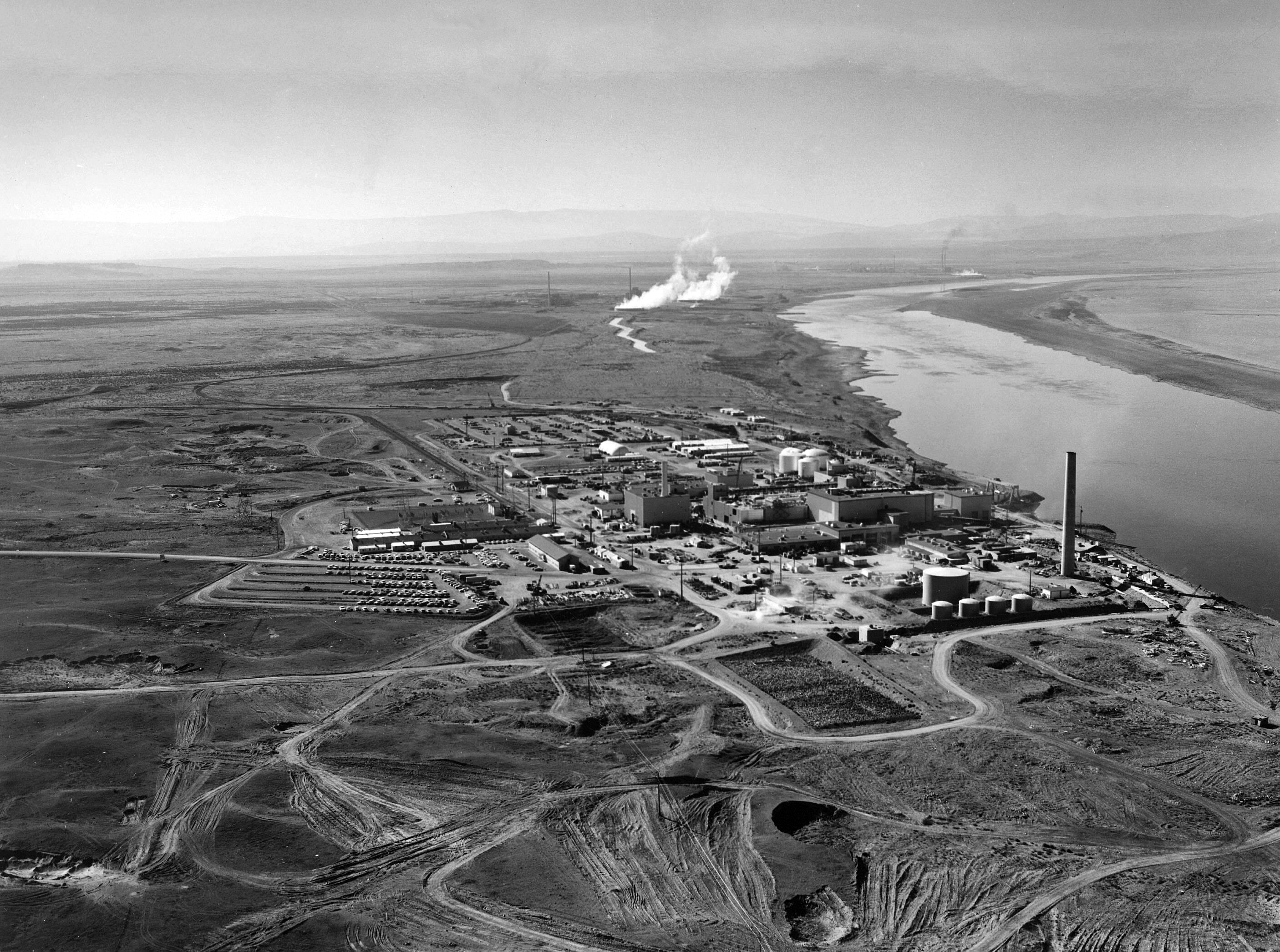 A black and white aerial photo showing the reactor complex by the Columbia River. The land surrounding the site has been dug up and is showing the muddy top soil. In the distance, near the water's edge are plumes of steam rising from buildings fading into the distance. The land beyond the complex is largely rolling fields leading to mountains that are just about visible. 