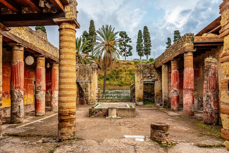 A photo taken within the square of a rich house building in Herculaneum. The square has pillars and overhanging roofs either side. Most of the pillars and the walls behind them have rich red paint while the others appear to be sandstone. Trees and a raised grassy bank are visible in the background.  