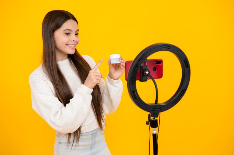 young girl holding a pot of skincare product, standing in front of a phone set up with a ring light, with a bright yellow background