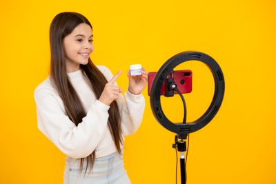young girl holding a pot of skincare product, standing in front of a phone set up with a ring light, with a bright yellow background