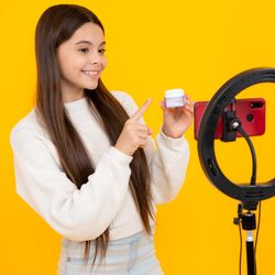 young girl holding a pot of skincare product, standing in front of a phone set up with a ring light, with a bright yellow background