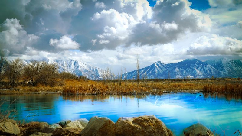 A photo taken by the side of Utah Lake, showing beautifully blue water in the foreground set against the mountains in the background. The sky is filled with thick clouds. 
