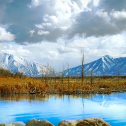 A photo taken by the side of Utah Lake, showing beautifully blue water in the foreground set against the mountains in the background. The sky is filled with thick clouds. 