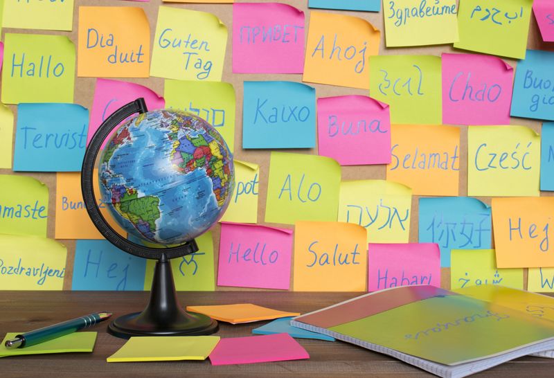 A photo showing a board with different colored post-it notes with "hello" written in different languages from around the world. There is a table in the bottom of the photo showing a small globe and some blank post-it note packs and a slim book.
