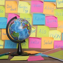 A photo showing a board with different colored post-it notes with "hello" written in different languages from around the world. There is a table in the bottom of the photo showing a small globe and some blank post-it note packs and a slim book.
