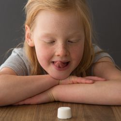 A young girl sits at a table across from a single marshmallow, attempting the marshmallow test