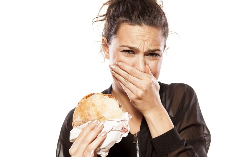 A woman covers her mouth and looks disgusted while she is holding a meat-filled burger. She is wearing a black leather jacket and her hair is tied up. 