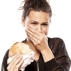 A woman covers her mouth and looks disgusted while she is holding a meat-filled burger. She is wearing a black leather jacket and her hair is tied up. 