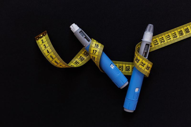 two blue and gray pens, such as those used to inject semaglutide, on a black background with a yellow tape measure entwined around them