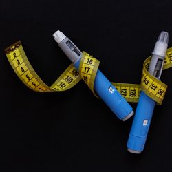 two blue and gray pens, such as those used to inject semaglutide, on a black background with a yellow tape measure entwined around them