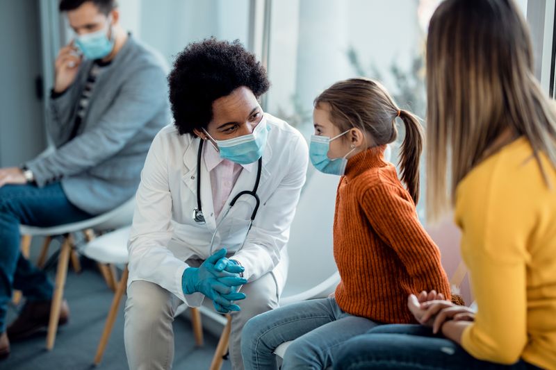 paediatrician wearing a white coat and a surgical masks speaking to a little girl also wearing a surgical mask in a waiting room, while her mother watches. There's a man in the background also waiting, wearing a mask.