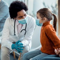 paediatrician wearing a white coat and a surgical masks speaking to a little girl also wearing a surgical mask in a waiting room, while her mother watches. There's a man in the background also waiting, wearing a mask.