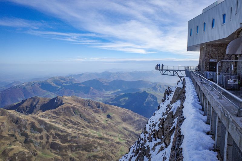 The photo shows a view point sticking out over the mountain side at Pic du Midi. The platform is in the distance from the camera's position and there are a few people standing at its end. It is position to the right hand side of the image and shows off the wide expanse of the Pyrenees mountains below it. The observatory itself is also partially visible to the extreme right of the shot.  