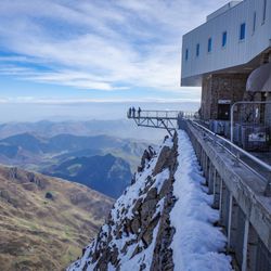 The photo shows a view point sticking out over the mountain side at Pic du Midi. The platform is in the distance from the camera's position and there are a few people standing at its end. It is position to the right hand side of the image and shows off the wide expanse of the Pyrenees mountains below it. The observatory itself is also partially visible to the extreme right of the shot.  
