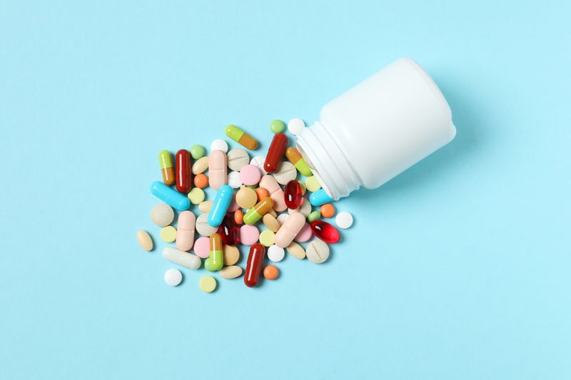 multicoloured pills and capsules spilling out of a plain white bottle on a pale blue background
