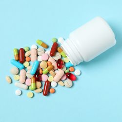multicoloured pills and capsules spilling out of a plain white bottle on a pale blue background