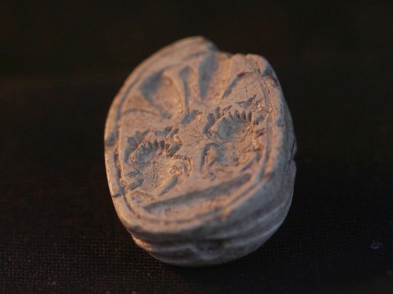 A close up photo of the reverse of the scarab seal showing its flatter side with a seal carved into it. There are two scarab shapes carved into it, along with some other impressions as decorations. It is possible to see the curved shape of the seal's reverse that is away from the camera.