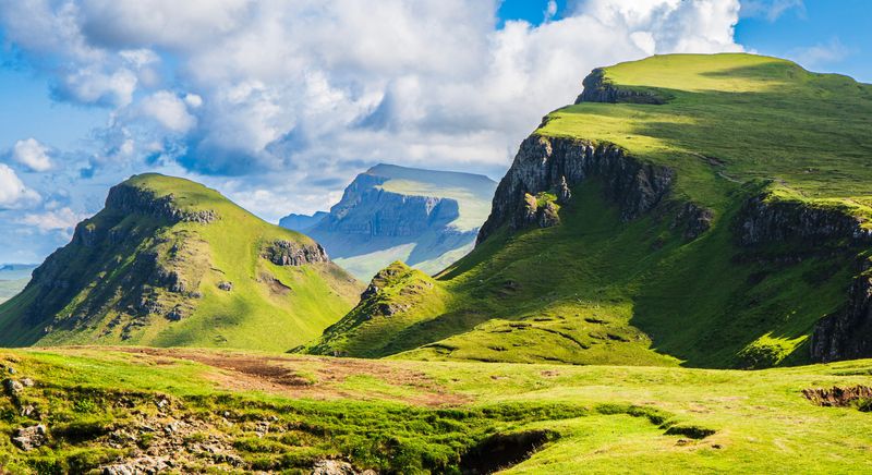 "Panoramic view of the Quiraing on the Isle of Skye, Scottish Highlands, with rolling green hills, steep cliffs, and dramatic shadows under a bright sky."
