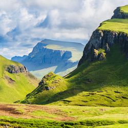 "Panoramic view of the Quiraing on the Isle of Skye, Scottish Highlands, with rolling green hills, steep cliffs, and dramatic shadows under a bright sky."