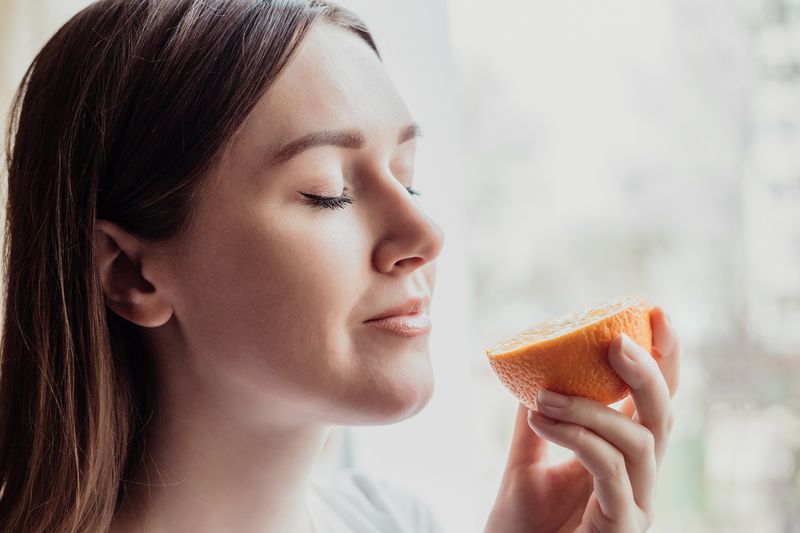 Close up portrait of young woman standing near a window and sniffing an orange