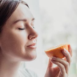 Close up portrait of young woman standing near a window and sniffing an orange