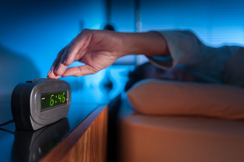 An arm reaching from under the bed covers to press snooze on a digital clock that says 6:45am
