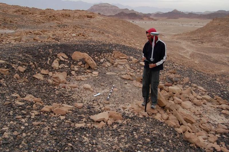A male researcher stands on a gravelly slope among loose stones. He is wearing a red hooded top and holding a large instrument that is as tall as he is. The slope is situated within a desert landscape of similarly coloured rocks and hillsides. 
