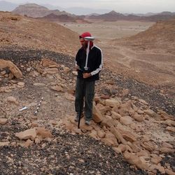 A male researcher stands on a gravelly slope among loose stones. He is wearing a red hooded top and holding a large instrument that is as tall as he is. The slope is situated within a desert landscape of similarly coloured rocks and hillsides. 