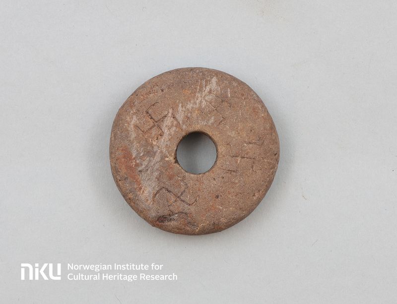 A photo showing the spinning whorl with the four swastikas carved into it. The object is rough and made of red sandstone and the symbols have been carved by hand in a crude manner. 