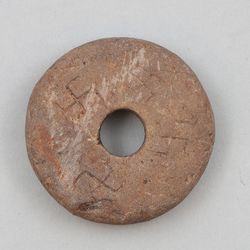 A photo showing the spinning whorl with the four swastikas carved into it. The object is rough and made of red sandstone and the symbols have been carved by hand in a crude manner. 