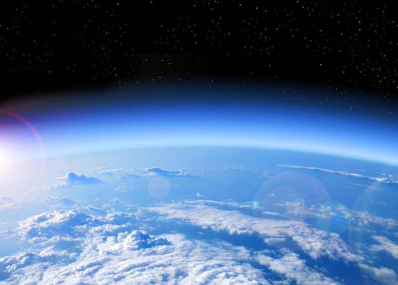 A photo taken just above the Earth's stratosphere, showing thick cloud cover stretching out to the curvature of the planet in the lower half of the image. The sky is blue and framed against the deep black of space above it. To the left of the photo, at the horizon point, a bright flare can be seen as the Sun is starting to rise into view. 