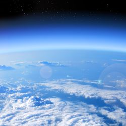 A photo taken just above the Earth's stratosphere, showing thick cloud cover stretching out to the curvature of the planet in the lower half of the image. The sky is blue and framed against the deep black of space above it. To the left of the photo, at the horizon point, a bright flare can be seen as the Sun is starting to rise into view. 