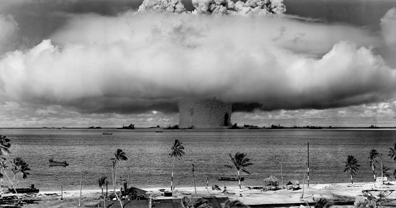 A black and white photo showing the enormous mushroom cloud caused by the nuclear explosion. The photo was taken from a distant island shoreline, the sandy beach of which is visible in the foreground. The mushroom cloud is set on the horizon and takes up most of the frame. 